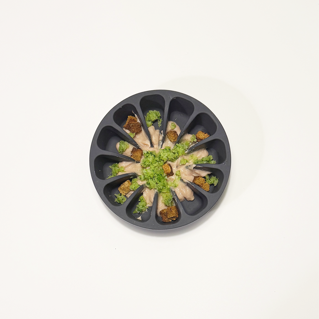 A dark gray circular silicone slow feeder filled with pureed broccoli, ham baby food and dried turkey treats